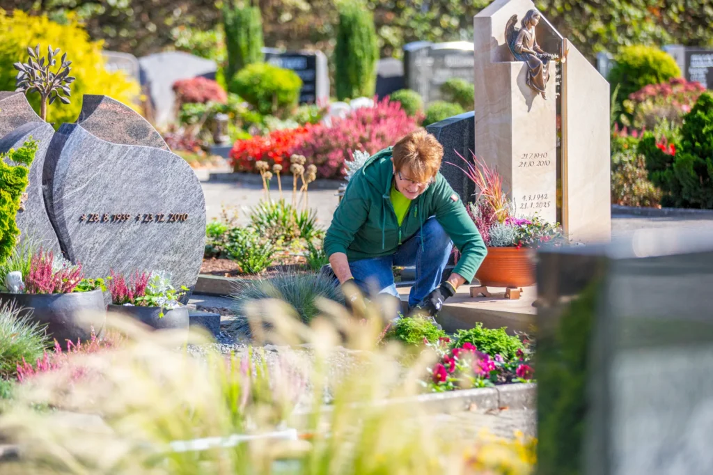 Frau mit braunen Haaren und grüner Jacke pflegt Gräber auf einem Friedhof, umgeben von bunten Blumen und Grabsteinen. Die Szenerie zeigt eine ruhige und gepflegte Umgebung, ideal für Gedenken und Trauer.