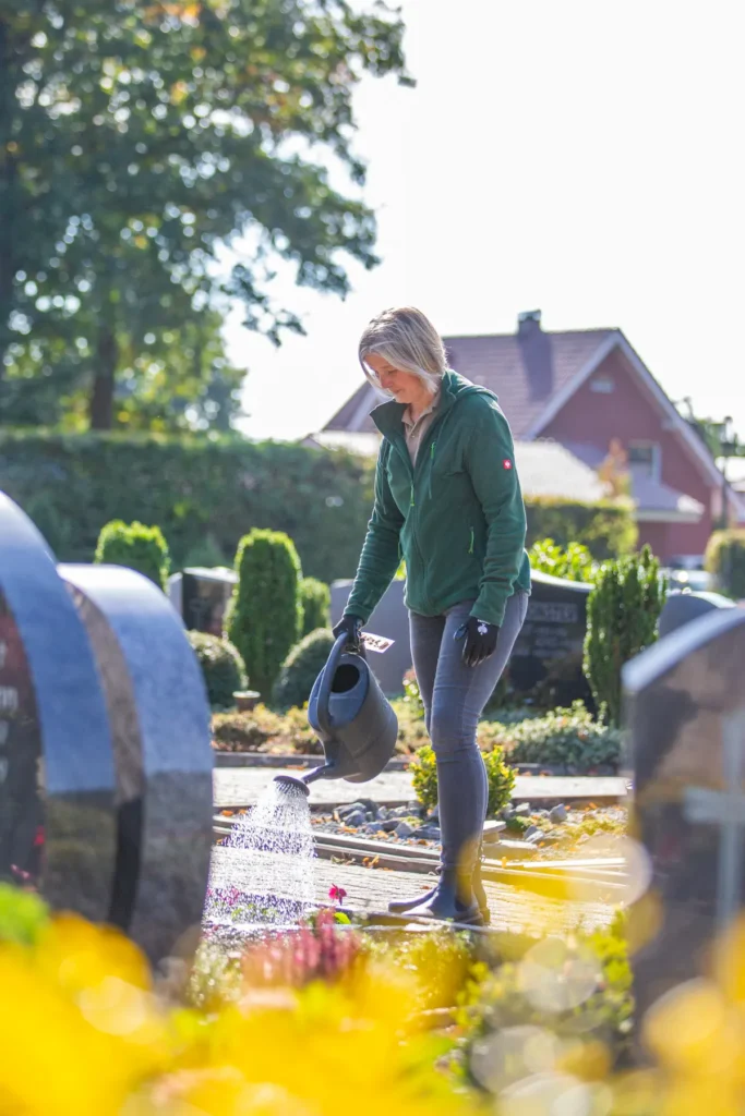 Eine Frau gießt Pflanzen auf einem Friedhof, umgeben von Grabsteinen und grünen Sträuchern, an einem sonnigen Tag.