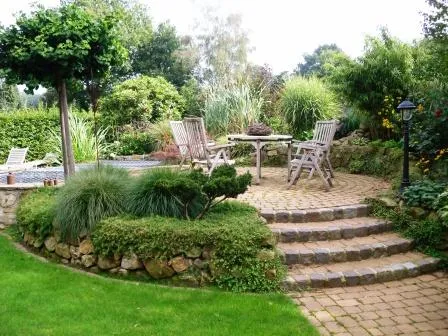 Ein schöner Garten mit einer Steinterrasse, zwei Holzstühlen und einem Tisch, umgeben von üppigem Grün und Blumen.