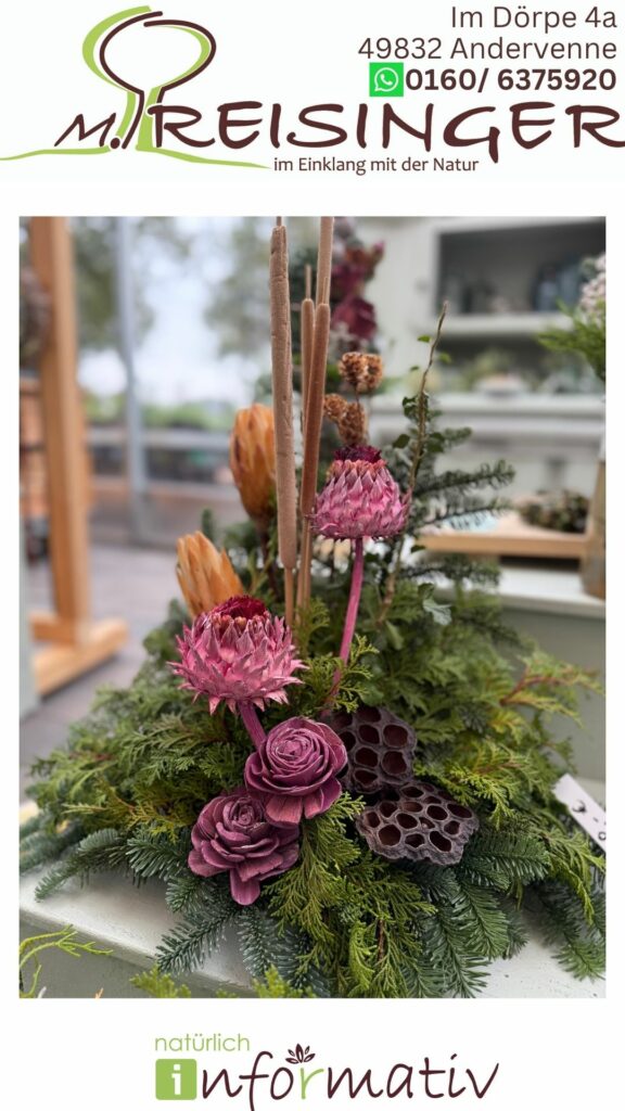 Ein florales Arrangement mit verschiedenen getrockneten Blüten, darunter pinkfarbene Protea, rosafarbene Rosen und natürliche Pflanzenmaterialien, umgeben von grünem Nadelgehölz. Im Hintergrund ist eine unscharfe Einrichtung sichtbar, die ein Einkaufslokal andeutet.