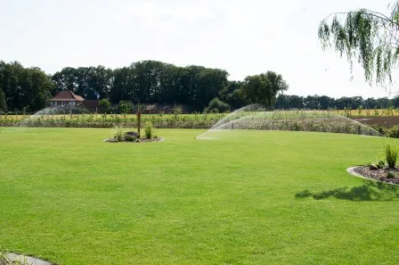 Grüner, gepflegter Rasen mit automatischen Sprinklern im Einsatz, umgeben von einem Garten mit Blumenbeeten und Bäumen, im Hintergrund eine ländliche Landschaft mit einem Wohnhaus und Wald.