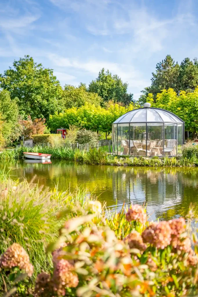 Ein idyllischer Garten mit einem gläsernen Gartenhaus am Ufer eines ruhigen Teichs, umgeben von üppigem Grün und bunten Blumen. Eine kleine, weiße Boot liegt sanft auf dem Wasser, während die Sonne den klaren Himmel erleuchtet und eine entspannte Atmosphäre schafft.