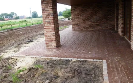 Terrasse mit roten Pflastersteinen und einem Backsteinsäule, umgeben von unbefestigtem Gelände und Wiesen im Hintergrund. Die Oberfläche ist sauber und neu, während der Bereich neben der Terrasse noch in der Bearbeitung ist.