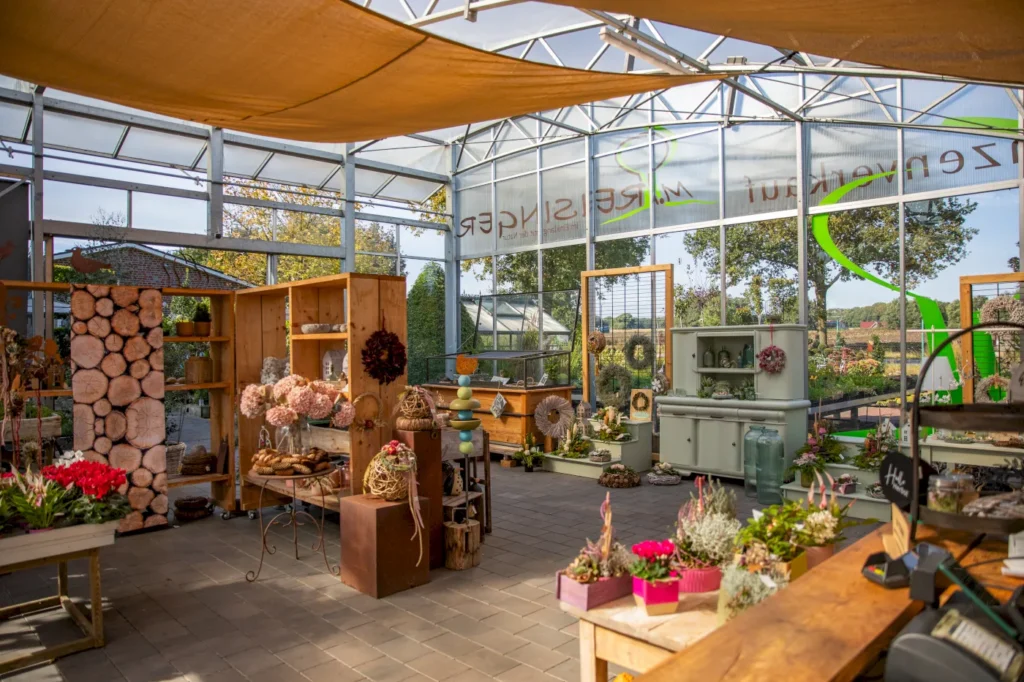 Blumenladen mit stilvollen Dekorationen und Arrangementen, darunter Holzregale mit Trockenblumen, Kränzen und Pflanzen. Die helle, glasüberdachte Umgebung bietet einen Blick auf die grüne Landschaft im Hintergrund. Verschiedene dekorative Elemente und Vasen sind auf den Tischen angeordnet, während die warmen Farben der Einrichtung eine einladende Atmosphäre schaffen.