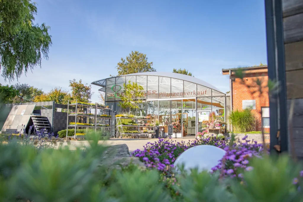 Moderne Verkaufsfläche für Pflanzen mit gläserner Fassade und einem begrünten Außenbereich. Vor dem Eingang sind bunte Blumen und Pflanzenarrangements zu sehen. Der Himmel ist blau und die Umgebung wirkt einladend und freundlich.