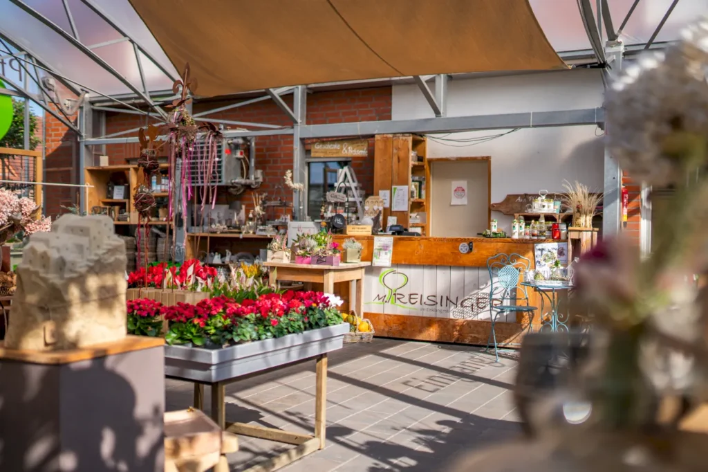 Blumenladen mit einer Vielzahl von bunten Pflanzen und Blumen in Töpfen, einem rustikalen Holztresen im Hintergrund, und dekorativen Elementen wie Skulpturen und Vasen. Der Raum ist hell erleuchtet und bietet eine einladende Atmosphäre für Kunden.