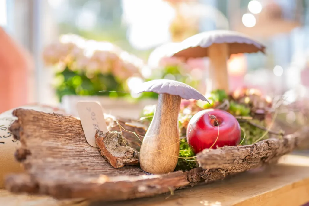 Bunte Pilze auf einem Holzbrett, umgeben von frischem Grün und einem roten Apfel, schaffen eine ansprechende, natürliche Stillleben-Atmosphäre.
