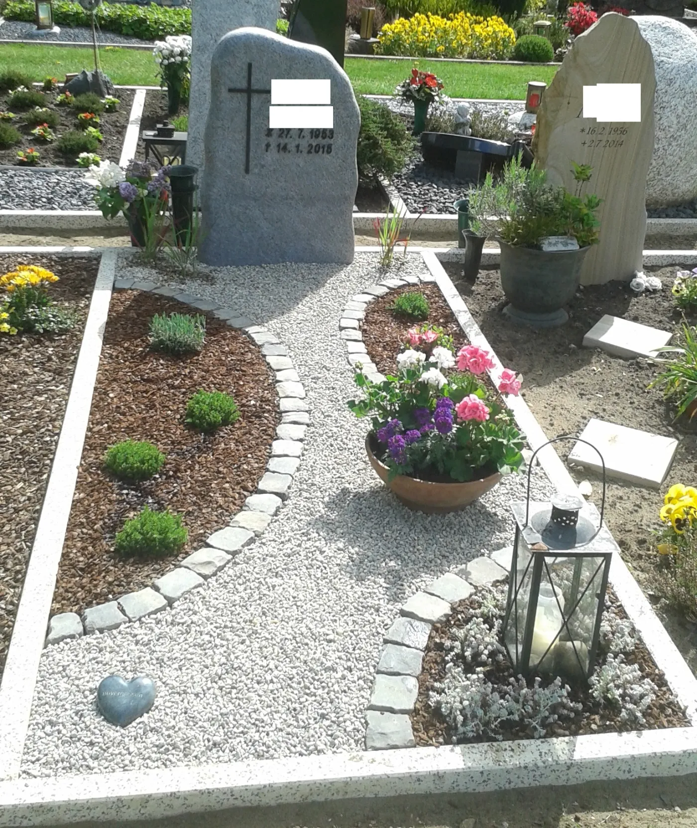 Grabstätte mit zwei Steinen, einer aus grauem und einer aus hellem Naturstein, umgeben von bunten Blumenbeeten und dekorativen Elementen wie einer Laterne und einem herzförmigen Stein. Der Weg ist mit Kies und kleinen Steinen gestaltet, während die Umgebung mit frischen Pflanzen und Blumen liebevoll gepflegt ist.