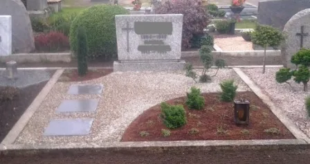 Gräberfeld mit mehreren Grabsteinen, umgeben von gepflegten Pflanzen und Kiesflächen. Der zentrale Grabstein ist aus grauem Stein und von einer harmonischen Anordnung kleiner Sträucher und Bäume umgeben. Eine dekorative Laterne steht auf dem Boden, während die Umgebung ruhig und gepflegt wirkt.