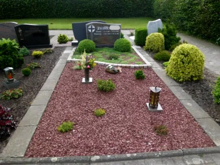 Friedhof mit Grabsteinen und gepflegtem Grabbereich, umgeben von bunten Sträuchern und Dekorationen.