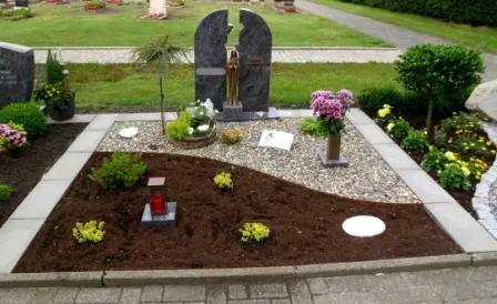 Grabstätte mit einer stilvollen Gestaltung, die eine zentrale Statue, bunte Blumenarrangements und verschiedene Pflanzen umfasst. Der Bereich ist mit Kies und frischem Erdboden versehen, umrahmt von grauen und weißen Platten. Die ruhige Umgebung wird durch gepflegte Grünflächen und einen klaren Weg ergänzt.