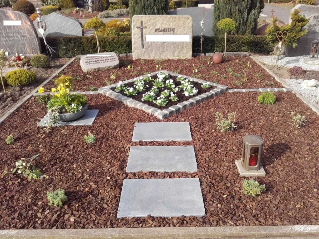 Das Bild zeigt eine liebevoll gestaltete Grabstätte, die durch sorgfältige Bepflanzung und ansprechende Gestaltung besticht. Im Zentrum der Fläche befindet sich ein kleiner Blumenbeet mit weißen und gelben Blüten, umrahmt von dekorativen Steinen. Die Verwendung von Rindenmulch sorgt nicht nur für eine ansprechende Optik, sondern auch für eine gute Bodenqualität und Feuchtigkeitsspeicherung. Die grauen Platten bieten einen klaren und stilvollen