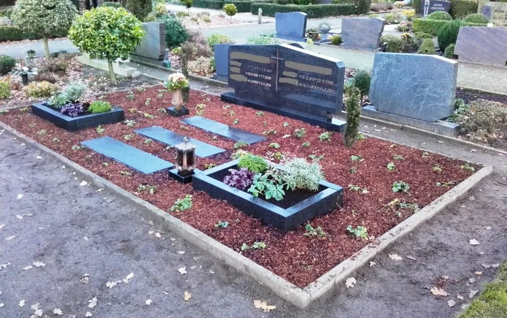 Grabstätte mit zwei schwarzen Grabsteinen, umgeben von frisch angelegten Blumenbeeten und dekorativem Mulch. Auf den Grabsteinen sind Inschriften zu sehen. Im Vordergrund befinden sich zwei Grablichter, und kleine Sträucher sowie Pflanzen ergänzen die Gestaltung des Grabes. Der Hintergrund zeigt weitere Grabsteine und eine gepflegte Friedhofsanlage.