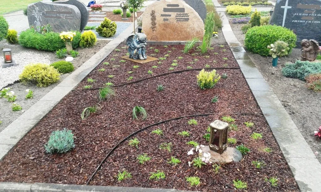 Das Bild zeigt eine liebevoll gestaltete Grabstätte, die mit einer Kombination aus Pflanzen und dekorativen Elementen versehen ist. Der Boden ist mit Rindenmulch bedeckt, was nicht nur ästhetisch ansprechend ist, sondern auch das Wachstum der Pflanzen fördert und Unkraut reduziert. In der Mitte der Grabstätte befindet sich eine kleine Statue, die einen trauernden Engel darstellt, was dem Ort eine besinnliche Atmosphäre verleiht. Um die Statue herum sind verschiedene