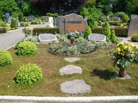 Ein gepflegter Friedhof mit einem Grabstein, umgeben von bunten Blumen und grünen Sträuchern auf blühendem Rasen.