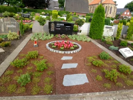 Gepflegtes Grab mit bunten Blumenbeeten, Kieswegen und grünen Sträuchern in einem ruhigen Friedhofsbereich.