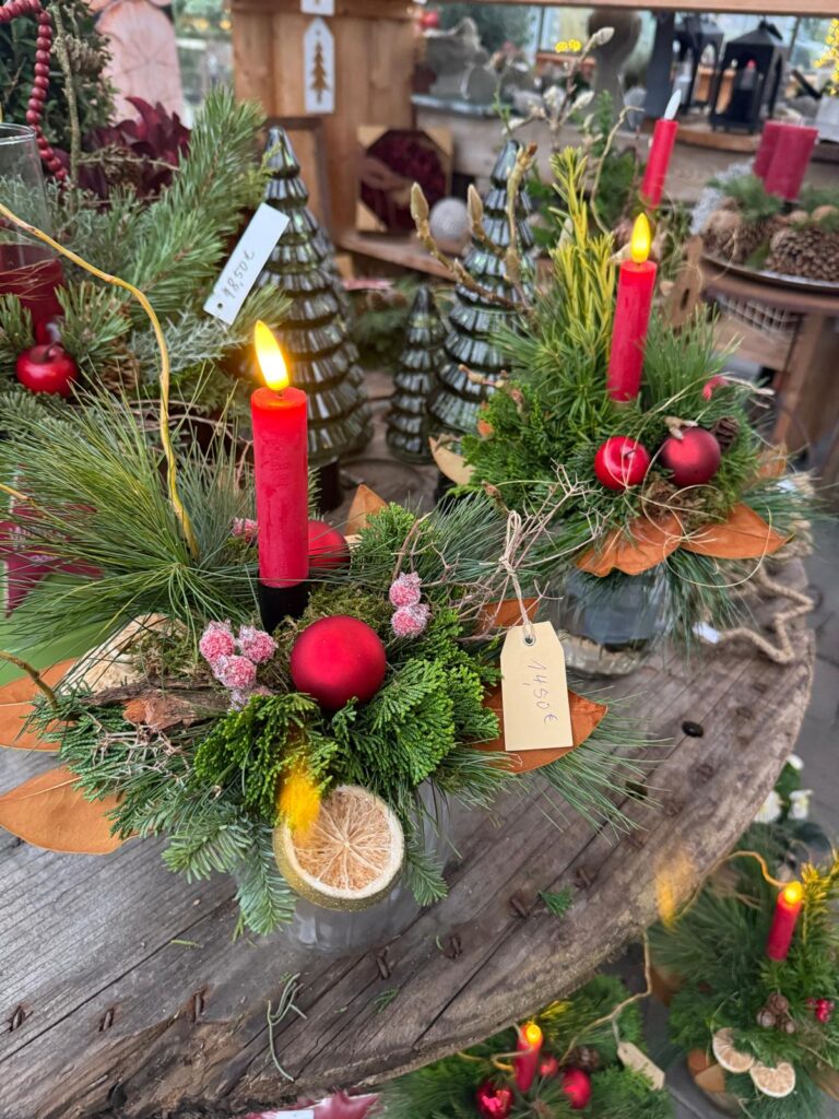 Ein weihnachtliches Arrangement mit roten Kerzen, dekorativen Äpfeln, Sukkulenten, verschiedenen Tannenzweigen und einer getrockneten Zitronenscheibe auf einem Holztablett, im Hintergrund stehen Weihnachtsbaum-Dekorationen.